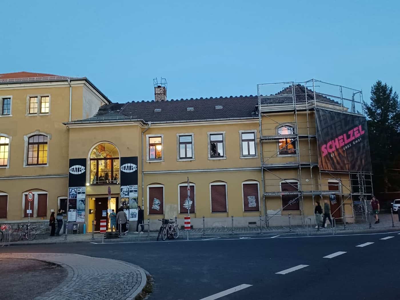 Gelbes, zweigeschossiges Gebäude des Konzertclubs Beatpol bei Dämmerung in Dresden-Briesnitz. Links ist der Eingang mit Plakaten und einem beleuchteten Bogenfenster zu sehen, die rechte Fassade ist teilweise eingerüstet.