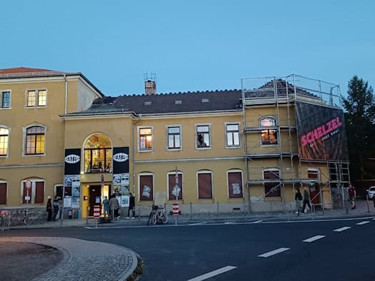 Gelbes, zweigeschossiges Gebäude des Konzertclubs Beatpol bei Dämmerung in Dresden-Briesnitz. Links ist der Eingang mit Plakaten und einem beleuchteten Bogenfenster zu sehen, die rechte Fassade ist teilweise eingerüstet.