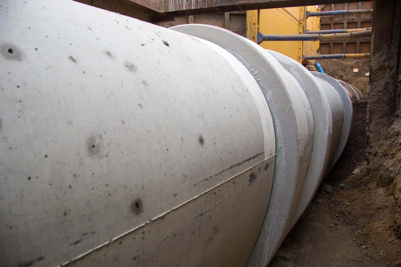 Blick in eine schmale Baugrube, in der mehrere massive Betonrohre in Reihe aneinandergefügt verlegt sind. Die Erdwand der Baugrube ist seitlich abgestützt.