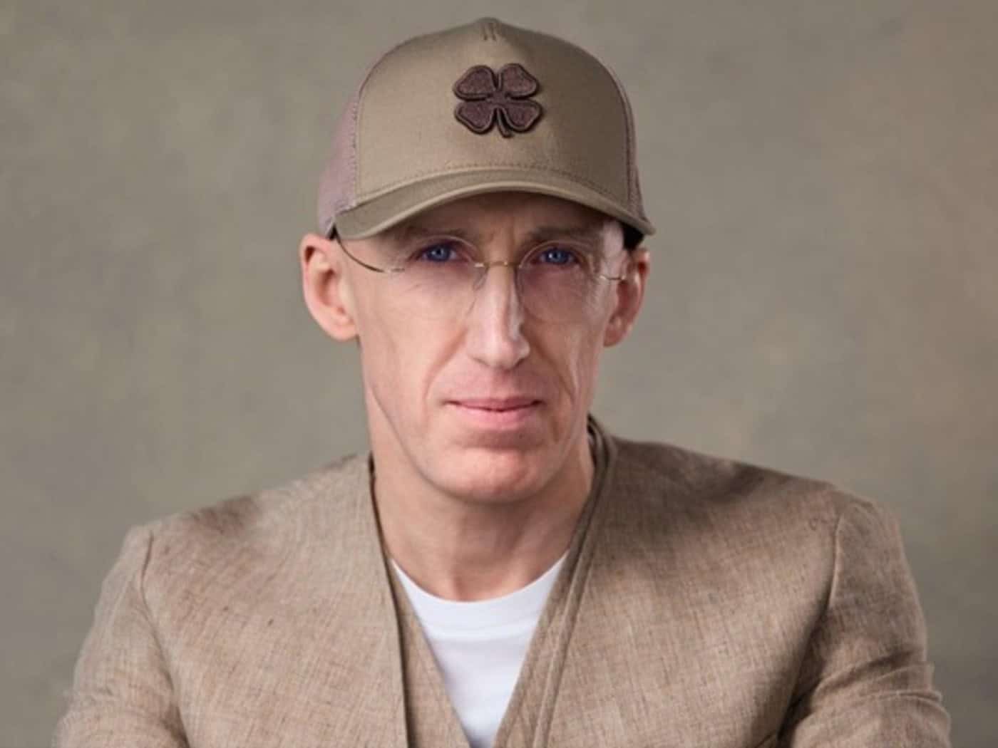 Porträt von Uri Poliavich mit Brille, beigefarbener Basecap und beigem Cardigan vor neutralem Hintergrund