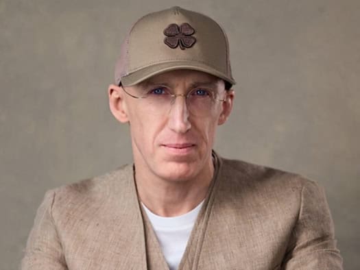 Porträt von Uri Poliavich mit Brille, beigefarbener Basecap und beigem Cardigan vor neutralem Hintergrund