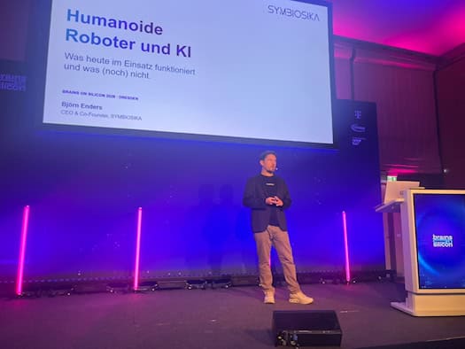 Björn Enders is standing next to a lectern on a stage lit in blue and purple. A large screen in the background displays a presentation slide with the heading “Humanoid Robots and AI” and the event name “Brains on Silicon Dresden.”