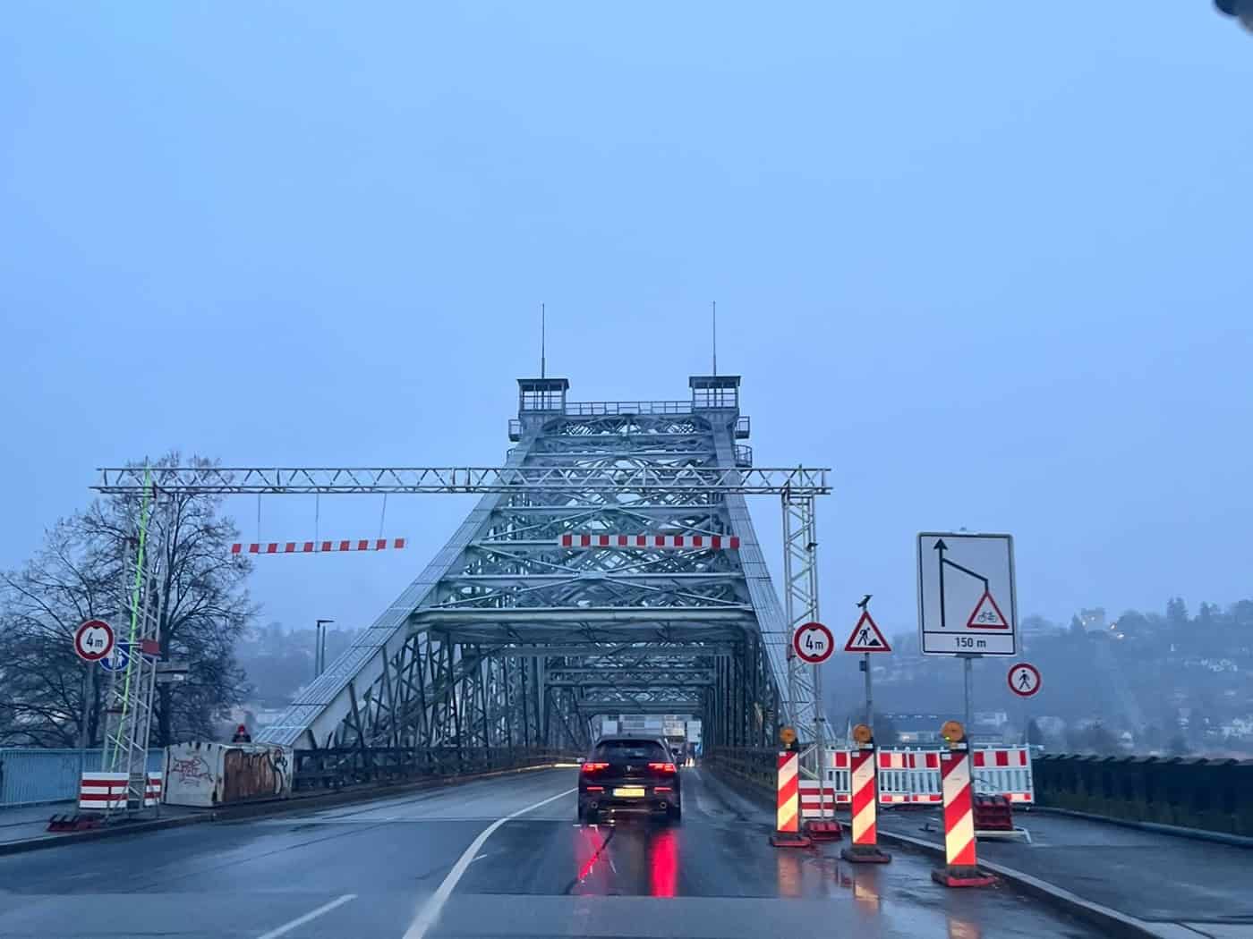 Blick aus der Fahrerperspektive eines Autos auf die nasse Fahrbahn der Loschwitzer Brücke (Blaues Wunder) in Dresden. Am Brückenkopf stehen Absperrbarken und Baustellenschilder, im Hintergrund erhebt sich das blaue Stahltragwerk bei trübem Wetter.