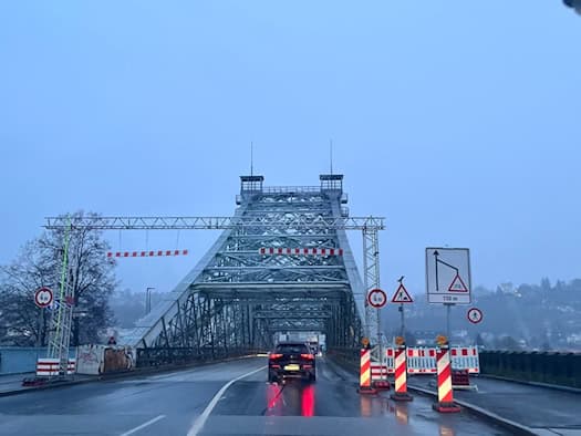 Blick aus der Fahrerperspektive eines Autos auf die nasse Fahrbahn der Loschwitzer Brücke (Blaues Wunder) in Dresden. Am Brückenkopf stehen Absperrbarken und Baustellenschilder, im Hintergrund erhebt sich das blaue Stahltragwerk bei trübem Wetter.