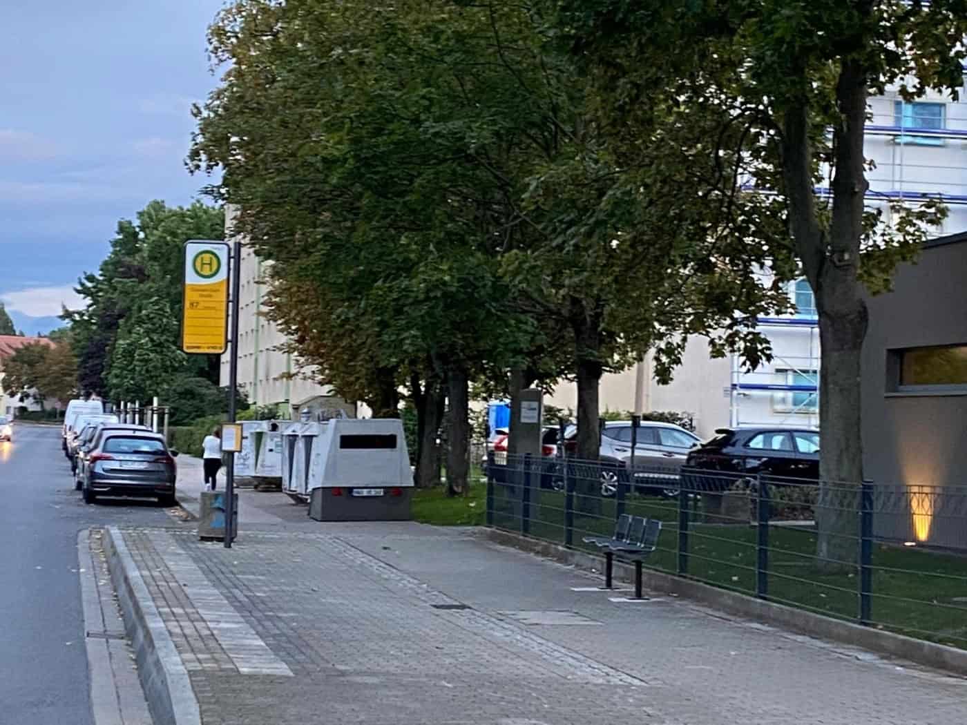 Ein grauer, kastenförmiger Blitzer-Anhänger steht auf dem Gehweg in Dresden-Leubnitz direkt neben einem gelben Bushaltestellenschild. Im Hintergrund stehen Bäume und geparkte Autos bei Dämmerung.