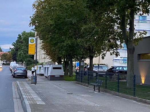 Ein grauer, kastenförmiger Blitzer-Anhänger steht auf dem Gehweg in Dresden-Leubnitz direkt neben einem gelben Bushaltestellenschild. Im Hintergrund stehen Bäume und geparkte Autos bei Dämmerung.