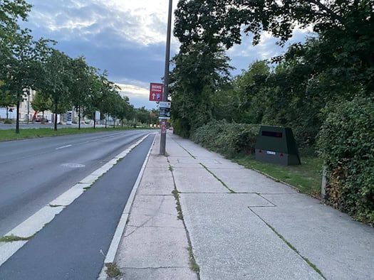 Straßenansicht des Zelleschen Wegs mit Gehweg und Radweg in Dresden. Am rechten Wegesrand steht vor grünem Gebüsch ein olivgrüner, trapezförmiger Blitzer-Anhänger mit dem Kennzeichen HWI-VE 213.