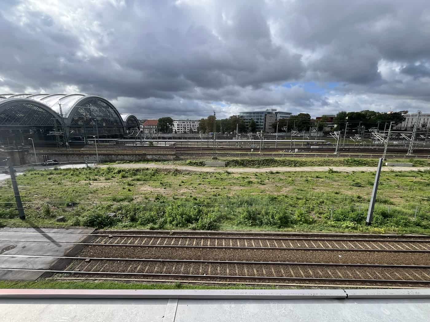 Weitwinkelaufnahme einer unbebauten, grün bewachsenen Brachfläche neben Eisenbahngleisen. Links ist die charakteristische Glaskuppeldach-Halle des Dresdner Hauptbahnhofs unter dynamischem Wolkenhimmel zu sehen.