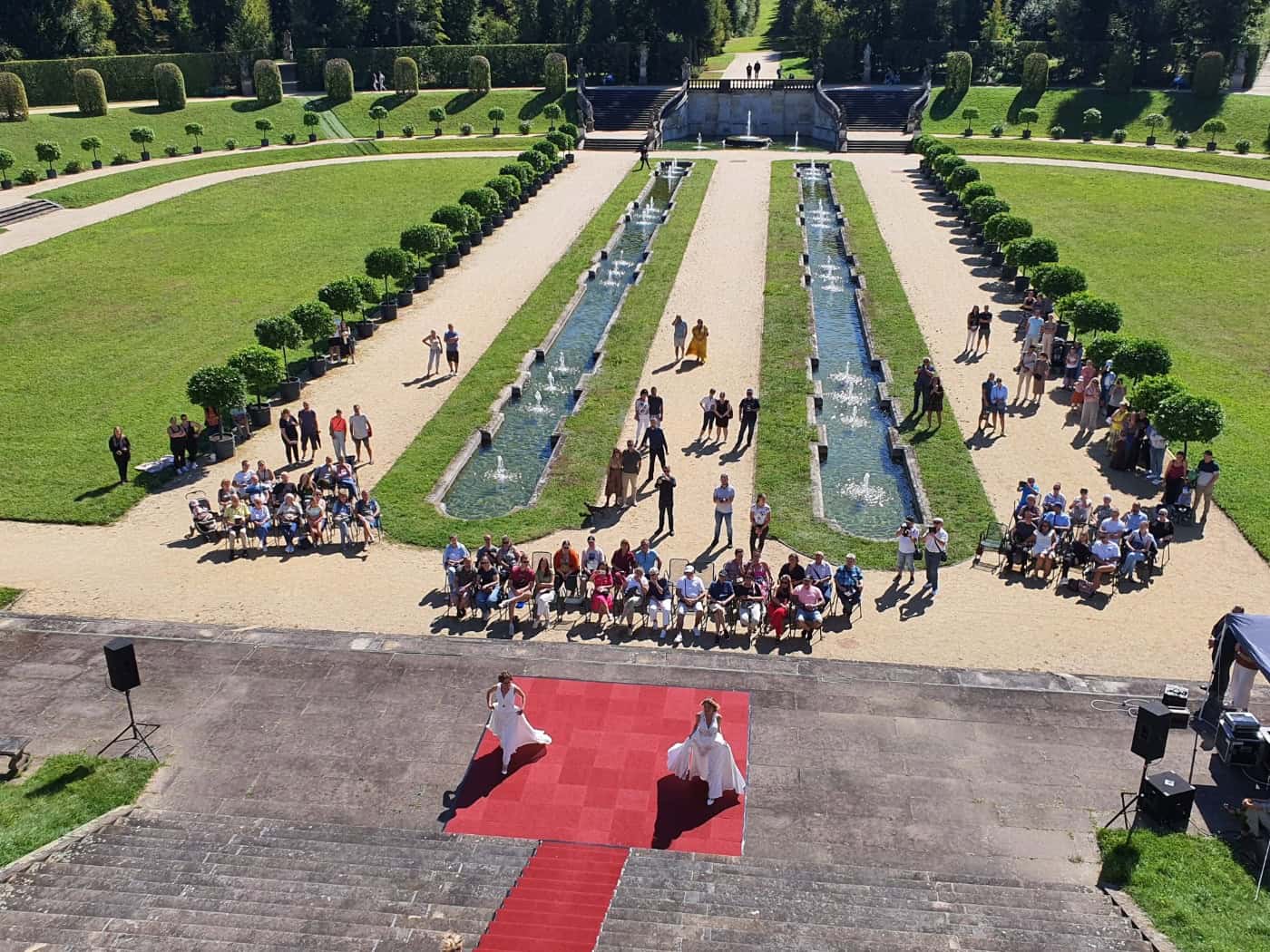 Blick von oben auf eine Freiluft-Brautmodenschau im Barockgarten Großsedlitz. Zwei Models in weißen Hochzeitskleidern laufen auf einem roten Teppich vor Zuschauern, die entlang symmetrischer Wasserbecken und Rasenflächen sitzen.
