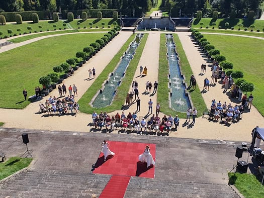 Blick von oben auf eine Freiluft-Brautmodenschau im Barockgarten Großsedlitz. Zwei Models in weißen Hochzeitskleidern laufen auf einem roten Teppich vor Zuschauern, die entlang symmetrischer Wasserbecken und Rasenflächen sitzen.