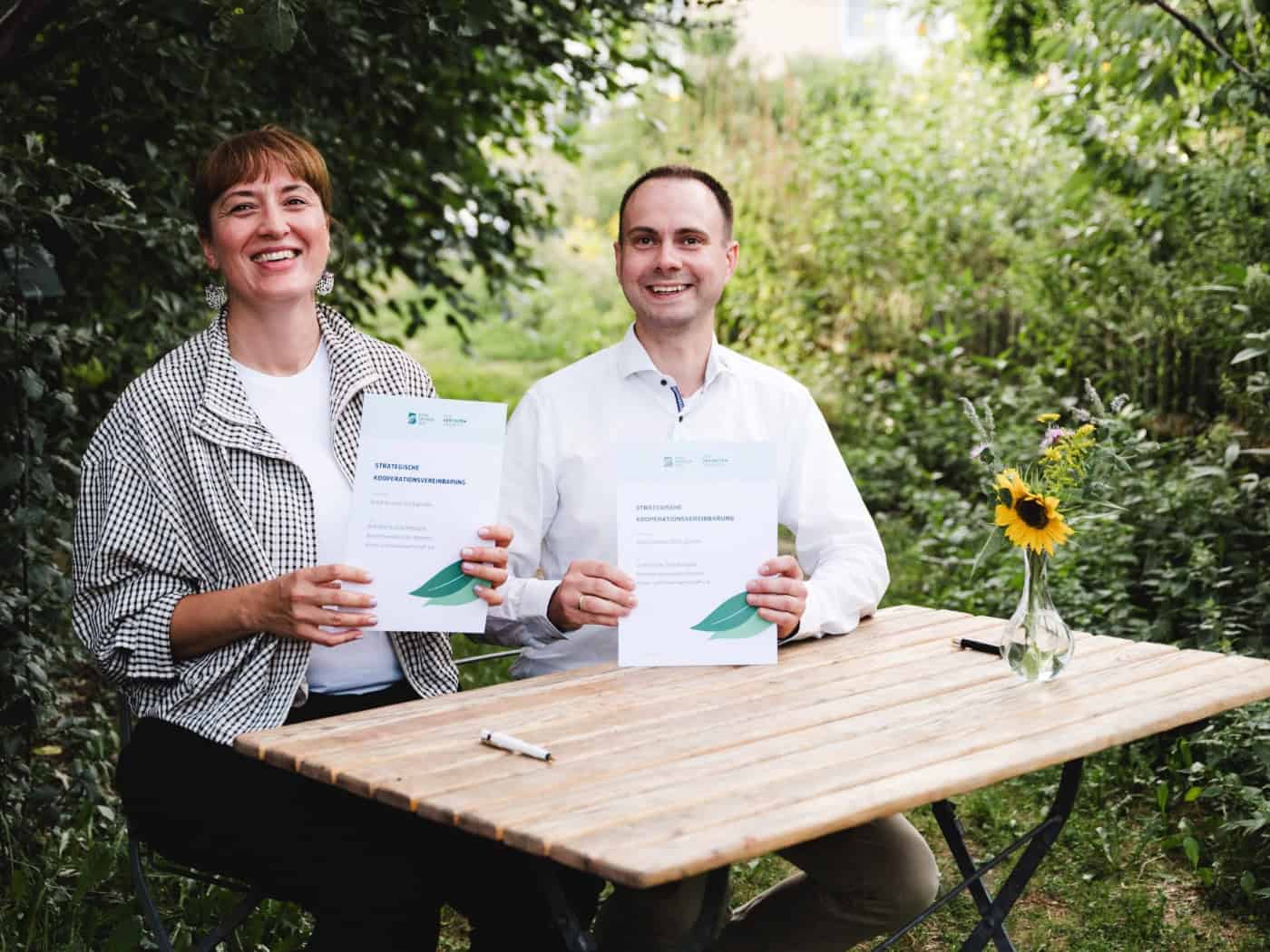 Marcel Timmroth und Frieda Pirnbaum sitzen lächelnd an einem Tisch im Freien vor grünem Hintergrund und unterzeichnen mit Stiften ein Dokument. Auf dem Tisch liegen Unterlagen und zwei Wassergläser.