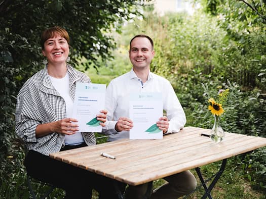 Marcel Timmroth und Frieda Pirnbaum sitzen lächelnd an einem Tisch im Freien vor grünem Hintergrund und unterzeichnen mit Stiften ein Dokument. Auf dem Tisch liegen Unterlagen und zwei Wassergläser.