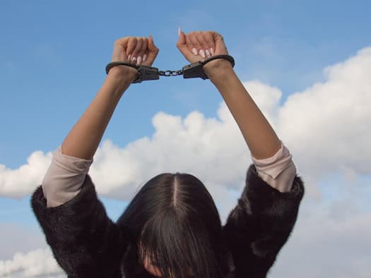 Symbolfoto: Eine dunkelhaarige Frau im dunklen Pelzmantel hält ihre gefesselten Hände mit schwarzen Handschellen vor blauem Himmel mit weißen Wolken in die Höhe. Der Kopf der Frau ist nach unten geneigt.