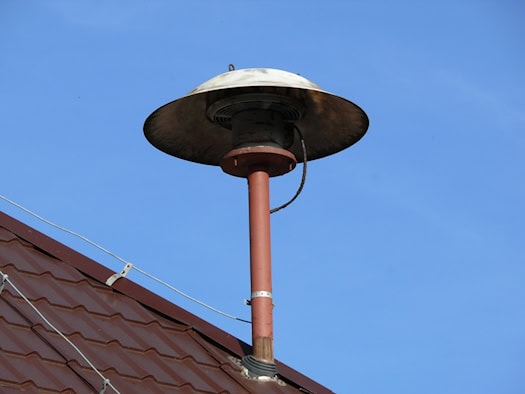 Eine Pilzsirene aus Metall ist auf dem braunen Ziegeldach eines Gebäudes montiert. Die alte Warnsirene hebt sich scharf von einem klaren, strahlend blauen Himmel ab.