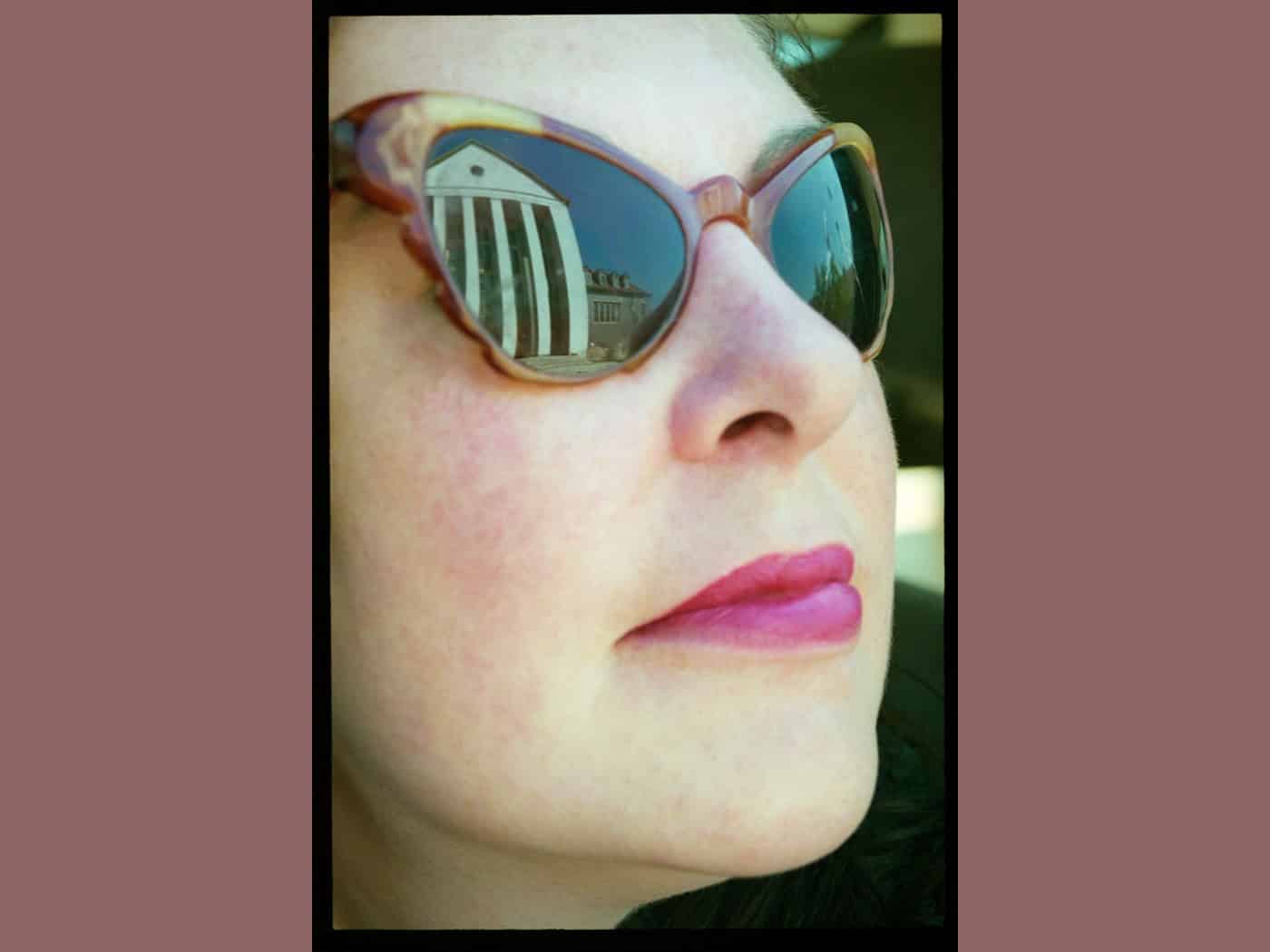 Nahaufnahme von Claudia Reichardt („Wanda“) von der Seite mit rotem Lippenstift und Cateye-Sonnenbrille. Im rechten Glas spiegelt sich die hellweiße Fassade des Festspielhauses Hellerau mit seinen dunklen Säulen unter blauem Himmel.