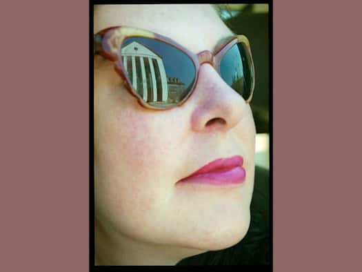 Nahaufnahme von Claudia Reichardt („Wanda“) von der Seite mit rotem Lippenstift und Cateye-Sonnenbrille. Im rechten Glas spiegelt sich die hellweiße Fassade des Festspielhauses Hellerau mit seinen dunklen Säulen unter blauem Himmel.