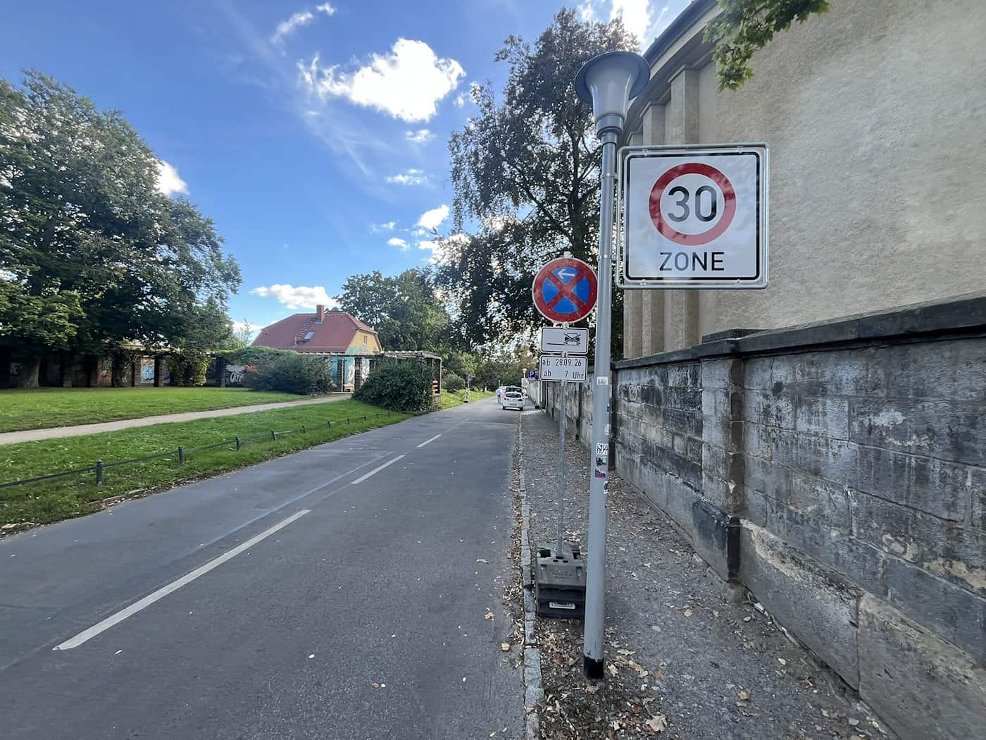 Ein Tempo-30-Zonen-Schild und ein absolutes Halteverbotsschild mit dem Zusatzschild „ab 28.09.26“ stehen an einer asphaltierten Straße neben einer alten Steinmauer.