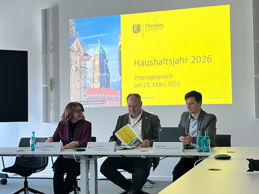 Oberbürgermeister Dirk Hilbert sitzt bei einer Pressekonferenz zwischen Jeanette Rißmann und Daniel Heine an einem Tisch. Im Hintergrund zeigt eine gelbe Leinwand die Aufschrift „Haushaltsjahr 2026 – Pressegespräch am 23. März 2026“.