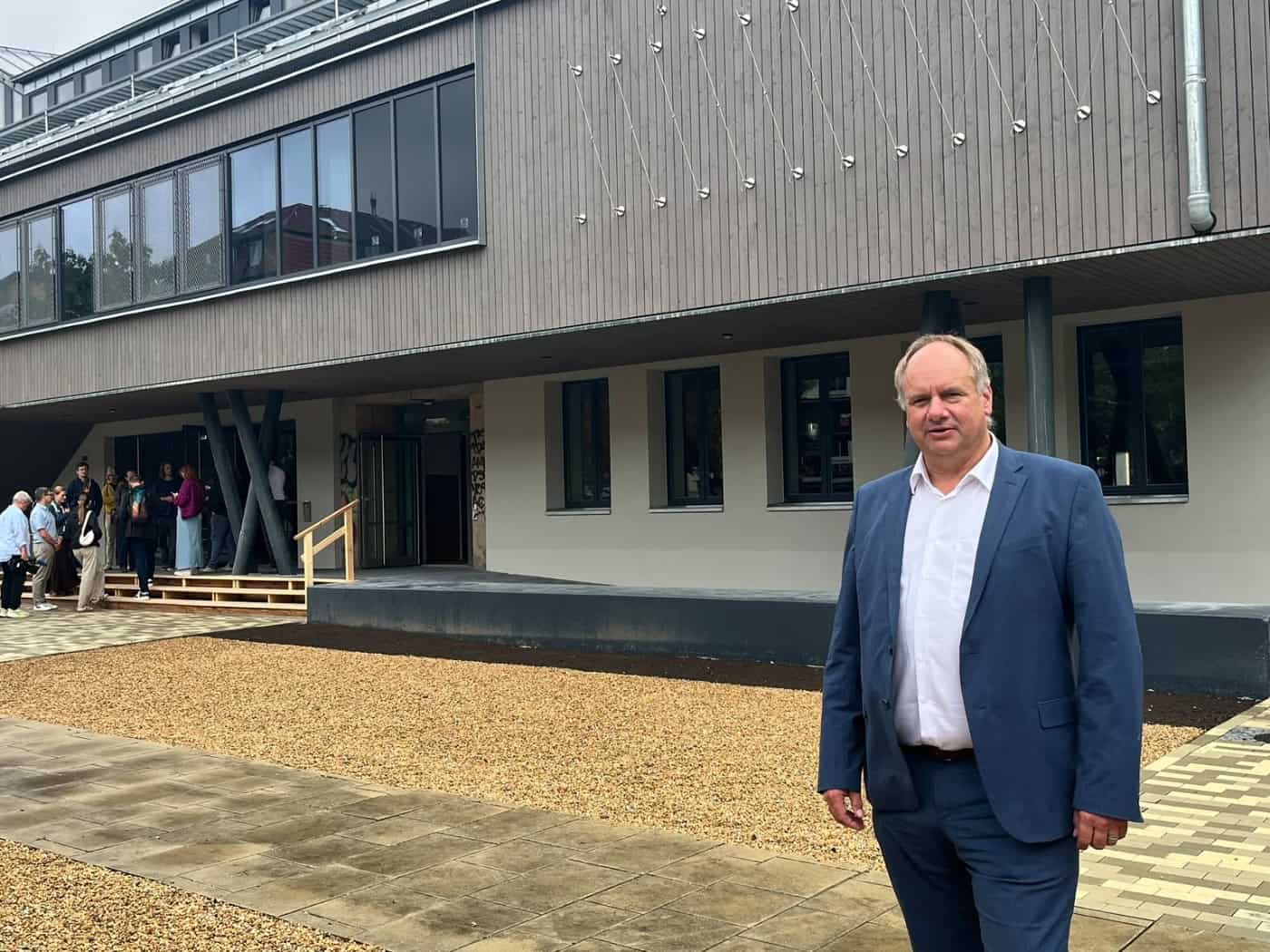 Dresdens Oberbürgermeister Dirk Hilbert steht im blauen Anzug auf dem geschotterten Vorplatz der neu gebauten Scheune. Im Hintergrund ist die Holzfassade des Kulturzentrums und der Eingang mit provisorischer Holztreppe zu sehen.