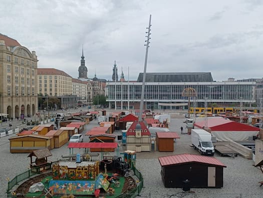 Blick von oben auf den Dresdner Altmarkt bei bewölktem Himmel. Auf dem Pflasterplatz stehen zahlreiche hölzerne Verkaufsbuden, Marktwagen und ein kleines Kinderkarussell im Aufbau. Im Hintergrund erstreckt sich die Glasfassade des Kulturpalastes.