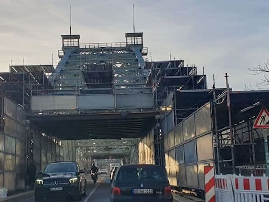 Blick auf die Fahrbahn des Blauen Wunders in Dresden, auf der Autos fahren. Das historische Stahlbauwerk und die Brückenpfeiler sind von einem gewaltigen Baugerüst mit Schutzverkleidungen überspannt.