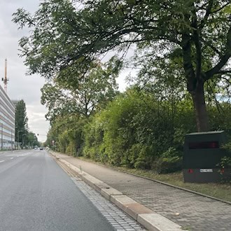 Blick aus der Perspektive eines Autofahrers auf die Strehlener Straße in Dresden: Am rechten Fahrbahnrand steht unter einem dichten Baum und vor einer Hecke der olivgrüne Messanhänger HWI-VE 213.