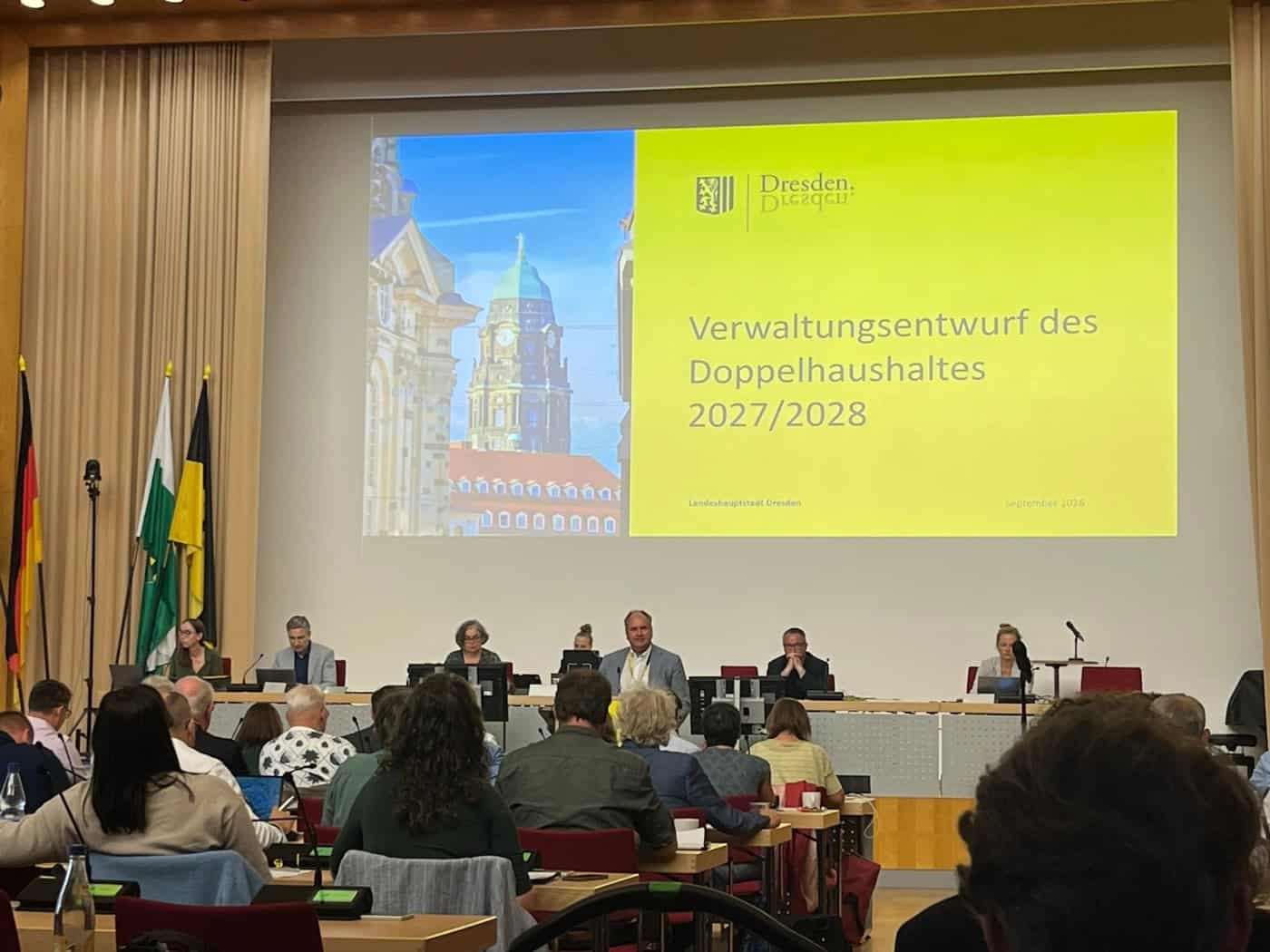 Große Leinwand in einem Tagungssaal zeigt den Titel „Verwaltungsentwurf des Doppelhaushaltes 2027/2028“. Vor der Projektion sitzt ein Podium mit Vertretern der Stadtverwaltung Dresden, im Vordergrund blickt das Publikum aus dem Saal auf die Bühne.