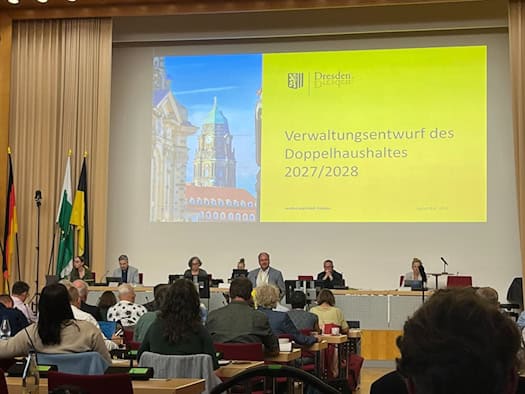 Große Leinwand in einem Tagungssaal zeigt den Titel „Verwaltungsentwurf des Doppelhaushaltes 2027/2028“. Vor der Projektion sitzt ein Podium mit Vertretern der Stadtverwaltung Dresden, im Vordergrund blickt das Publikum aus dem Saal auf die Bühne.