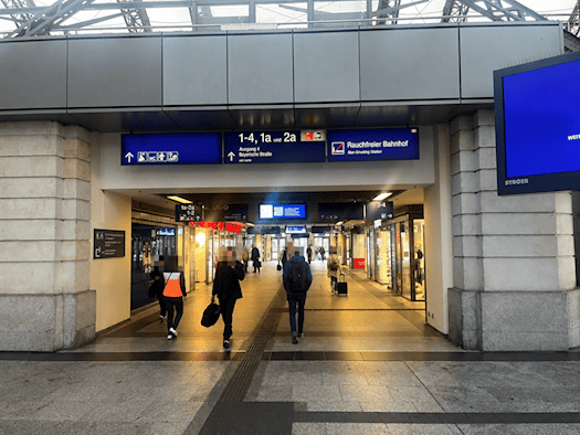 Blick in eine beleuchtete Passage im Dresdner Hauptbahnhof. Im Vordergrund gehen Reisende unter einem blauen Hinweisschild zu den Gleisen 1 bis 4 durch den Durchgang. Rechts hängt ein großer digitaler Bildschirm an der Wand.