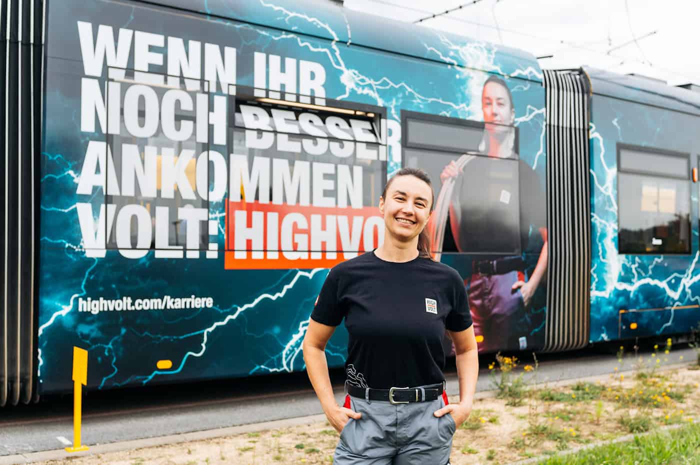 HIGHVOLT-Mitarbeiterin vor der neuen Kampagnen-Straßenbahn in Dresden Eine lächelnde Frau in schwarzem HIGHVOLT-T-Shirt und grauer Arbeitshose steht im Vordergrund. Direkt hinter ihr ist die Außenwand einer Straßenbahn zu sehen, die bedruckt ist mit der Aufschrift „WENN IHR NOCH BESSER ANKOMMEN VOLT: HIGHVOLT“ und ihrem Porträt.