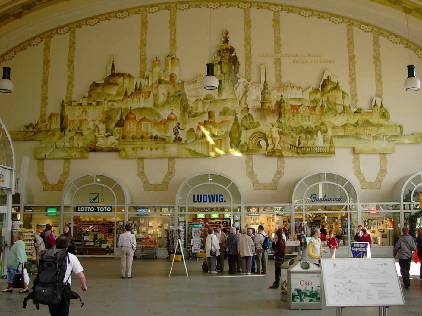 Die große Empfangshalle des Bahnhofs Dresden-Neustadt mit einem großen Wandgemälde historischer Schlösser an der gelben Wand. Darunter befinden sich Geschäfte wie Buch- und Tabakläden sowie Passanten mit Gepäck.