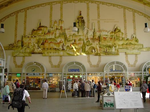 Die große Empfangshalle des Bahnhofs Dresden-Neustadt mit einem großen Wandgemälde historischer Schlösser an der gelben Wand. Darunter befinden sich Geschäfte wie Buch- und Tabakläden sowie Passanten mit Gepäck.
