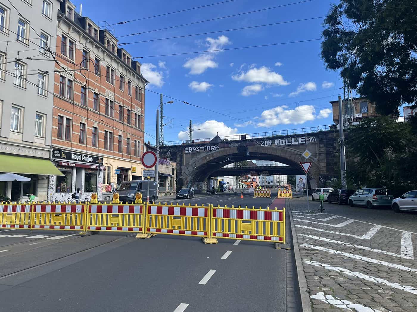 Rot-gelbe Absperrgitter blockieren die Fahrbahn des Bischofswegs in Dresden vor einer großen Eisenbahnbrücke mit Graffiti unter blauem Sommerhimmel.