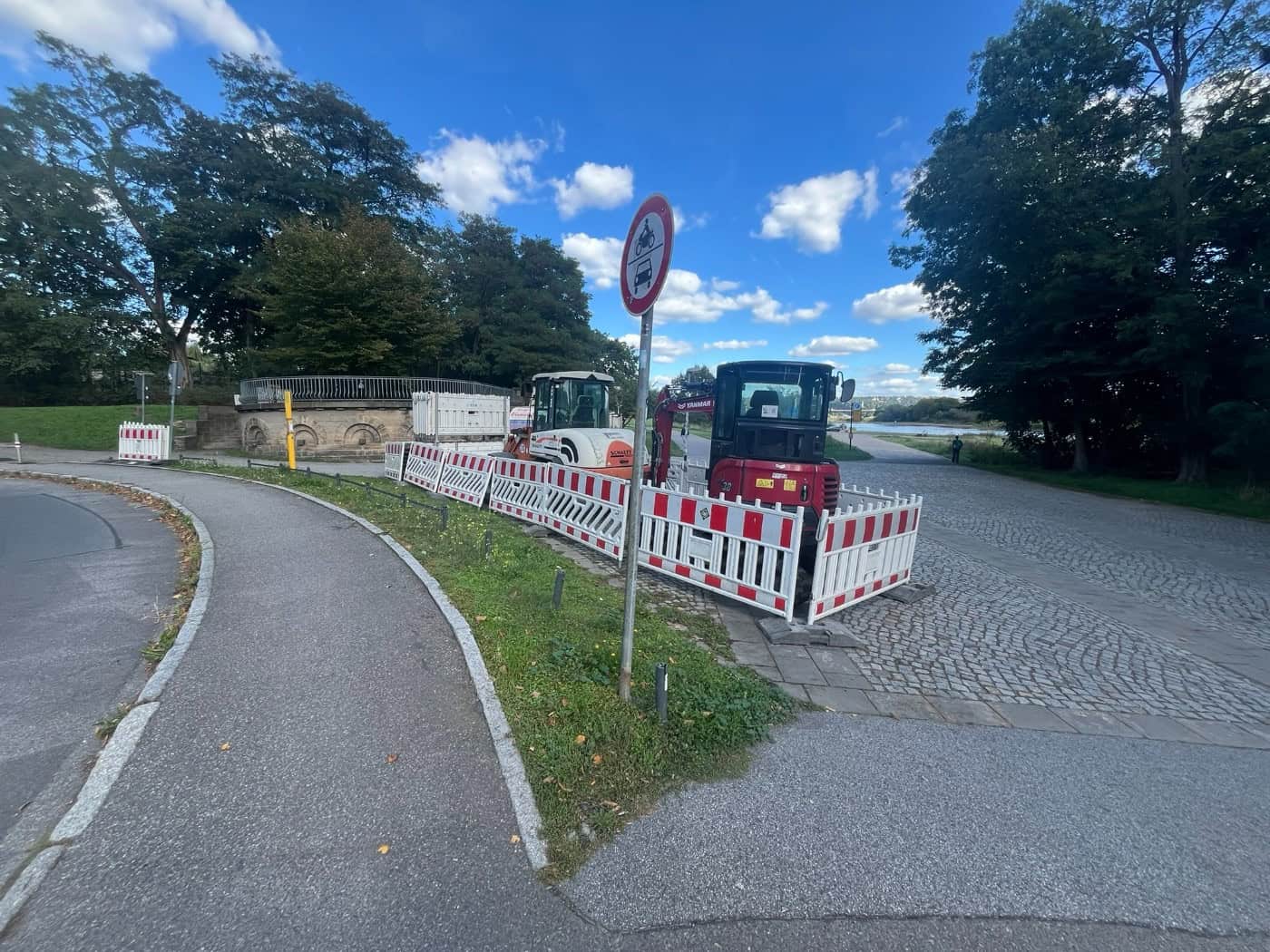 Roter Minibagger und Baustellenabsperrungen mit rot-weißen Zäunen am Diakonissenweg in Dresden neben einem Gehweg und Bäumen unter blauem Himmel mit Wolken.