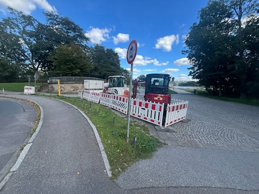 Roter Minibagger und Baustellenabsperrungen mit rot-weißen Zäunen am Diakonissenweg in Dresden neben einem Gehweg und Bäumen unter blauem Himmel mit Wolken.