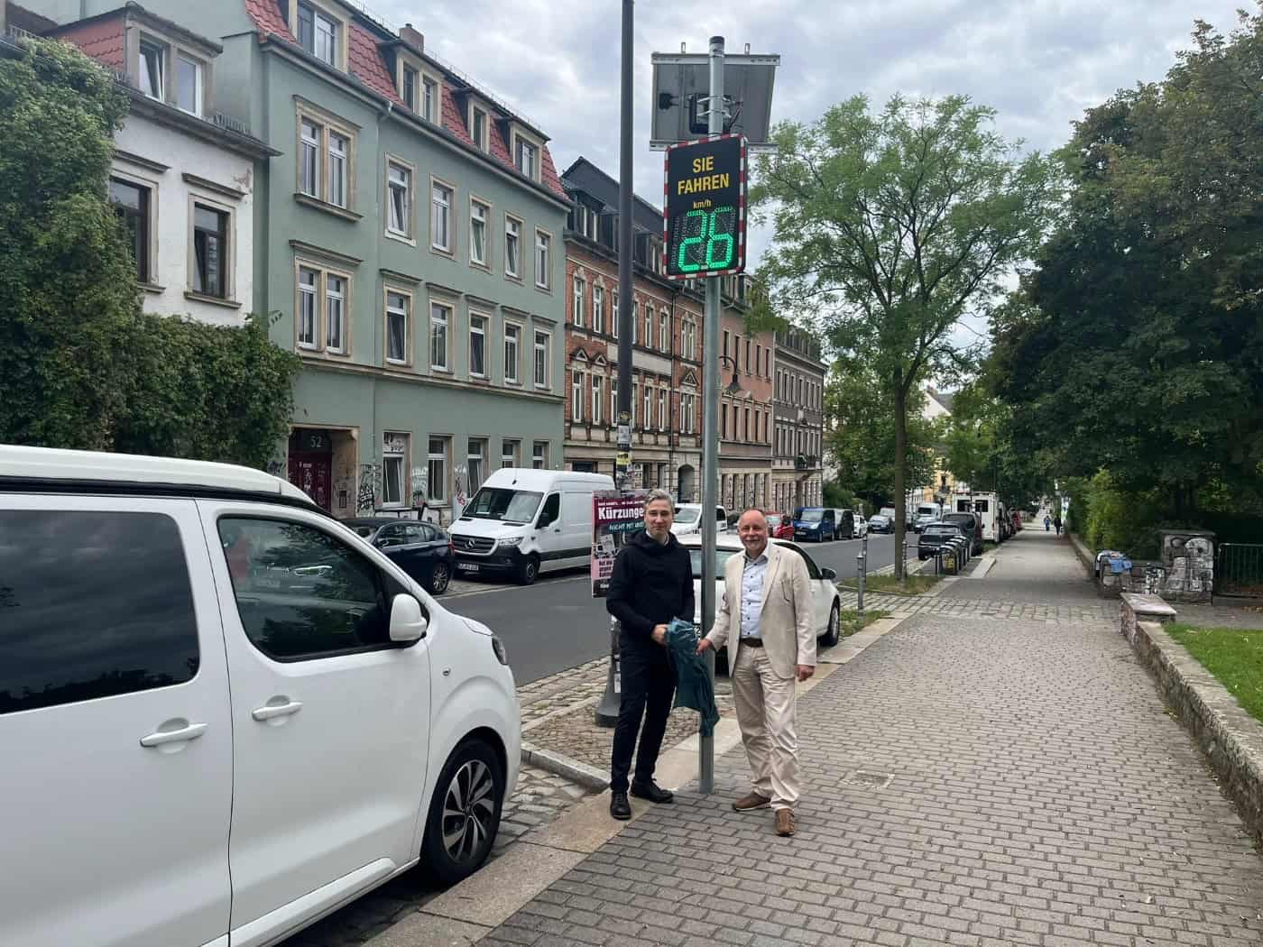 Stephan Kühn und André Barth stehen auf dem Gehweg der Rudolf-Leonhard-Straße neben einem Solar-Masten. Das digitale Display zeigt in grün „SIE FAHREN 26 km/h“ mit Smiley. Im Hintergrund stehen mehrstöckige Wohngebäude und geparkte Fahrzeuge.