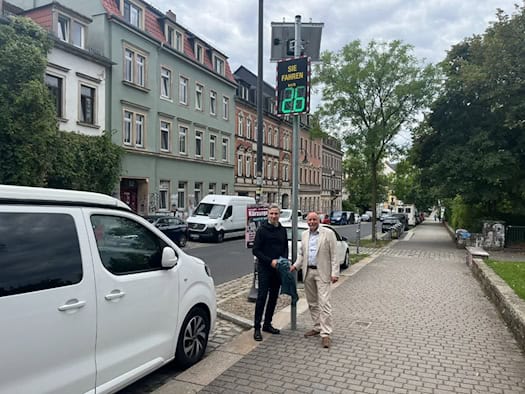 Stephan Kühn und André Barth stehen auf dem Gehweg der Rudolf-Leonhard-Straße neben einem Solar-Masten. Das digitale Display zeigt in grün „SIE FAHREN 26 km/h“ mit Smiley. Im Hintergrund stehen mehrstöckige Wohngebäude und geparkte Fahrzeuge.