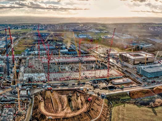 Luftaufnahme einer riesigen Großbaustelle für ein Chipwerk im Dresdner Norden. Zahlreiche rote und gelbe Baukräne stehen auf dem Areal mit Erdaushub und Rohbauten. Im Hintergrund sind angrenzende Felder und das Umland zu sehen.