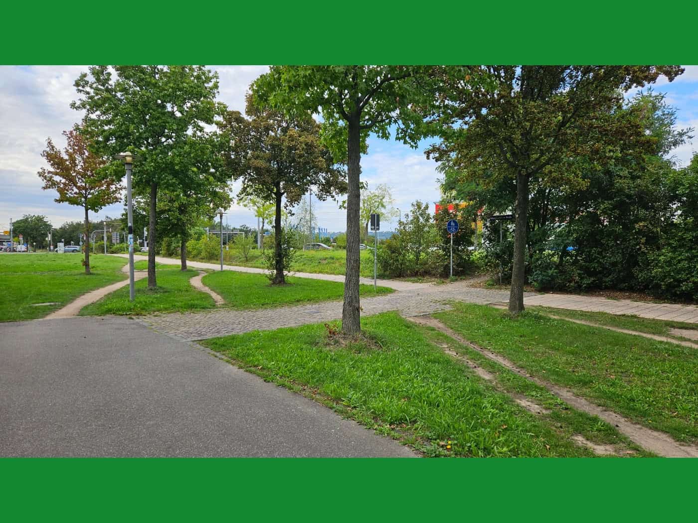 Ein asphaltierter Fuß- und Radweg führt an einer Grünfläche vorbei. Deutlich sichtbare, unbefestigte Trampelpfade wurden von Radfahrern quer über die Wiese gefahren, um den zickzackförmigen Pflasterweg abzukürzen. Bäume und Straßenschilder säumen das Areal.