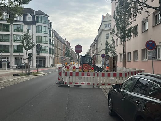 Straßenansicht in Dresden-Pieschen mit eingerichteter Baustelle: Rot-weiße Absperrgitter, ein kleiner orangefarbener Bagger und Verbotsschilder blockieren eine Fahrbahnseite der Rehefelder Straße zwischen mehrstöckigen Wohngebäuden.