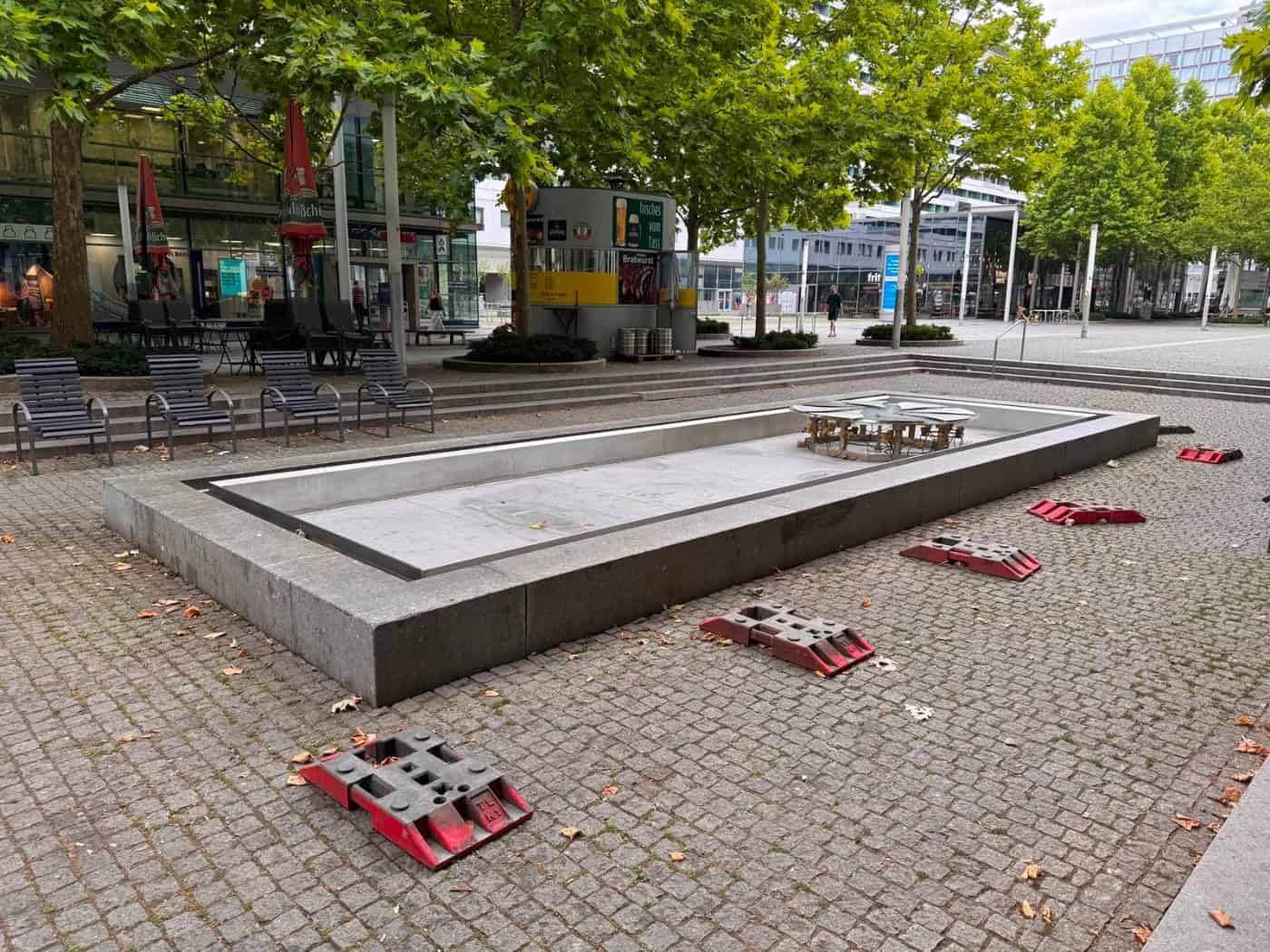 Das trockene, graue Steinbecken des Brunnens auf der gepflasterten Prager Straße in Dresden. Im leeren Becken ist die Brunnenmechanik zu sehen. Auf dem Pflaster davor liegen rote Fußplatten von Absperrungen, im Hintergrund 