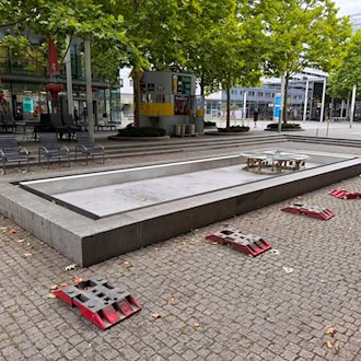 Das trockene, graue Steinbecken des Brunnens auf der gepflasterten Prager Straße in Dresden. Im leeren Becken ist die Brunnenmechanik zu sehen. Auf dem Pflaster davor liegen rote Fußplatten von Absperrungen, im Hintergrund 