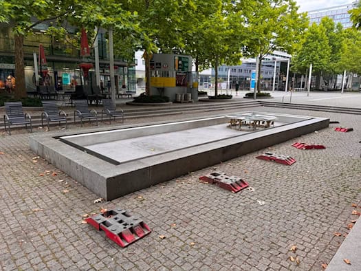 Das trockene, graue Steinbecken des Brunnens auf der gepflasterten Prager Straße in Dresden. Im leeren Becken ist die Brunnenmechanik zu sehen. Auf dem Pflaster davor liegen rote Fußplatten von Absperrungen, im Hintergrund 