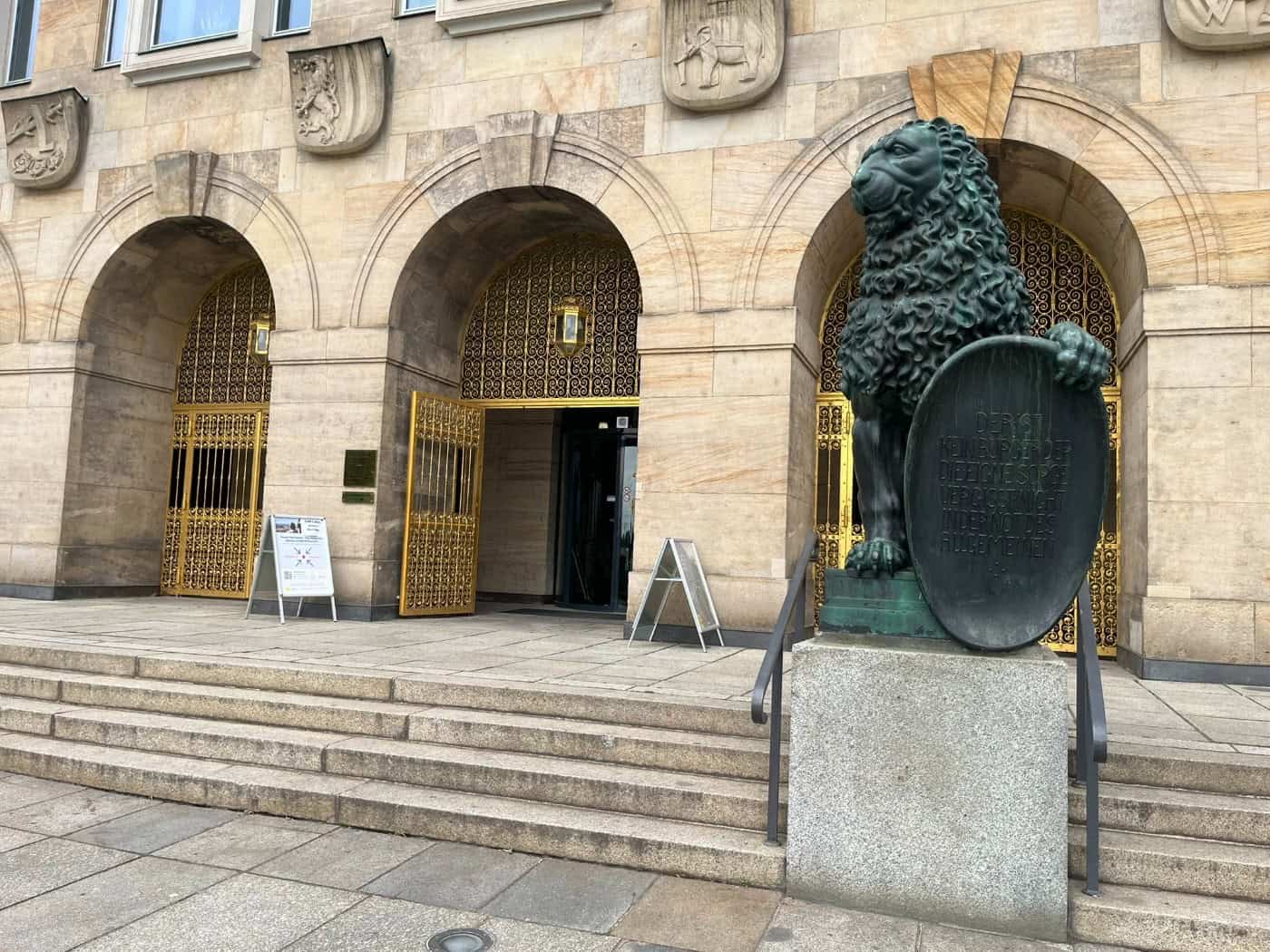 Haupteingang des Neuen Rathauses in Dresden mit breiter Treppe, Rundbögen und goldenen Verzierungen. Rechts im Vordergrund steht eine dunkle Bronzestatue eines sitzenden Löwen, der ein Schild hält.