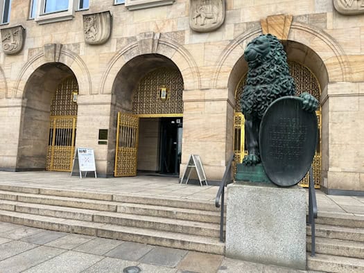 Haupteingang des Neuen Rathauses in Dresden mit breiter Treppe, Rundbögen und goldenen Verzierungen. Rechts im Vordergrund steht eine dunkle Bronzestatue eines sitzenden Löwen, der ein Schild hält.