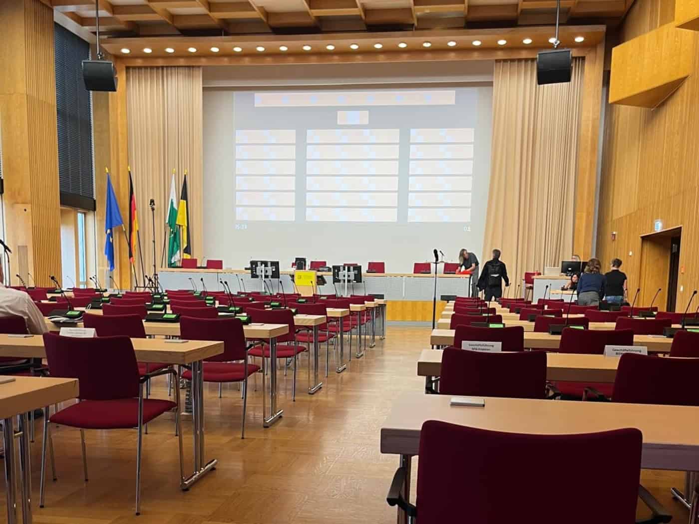 Blick durch die Reihen roter Stühle und Holztische im noch leeren Plenarsaal des Neuen Rathauses Dresden auf das Podium mit großer Leinwand und der Dresdner sowie sächsischen Flagge.