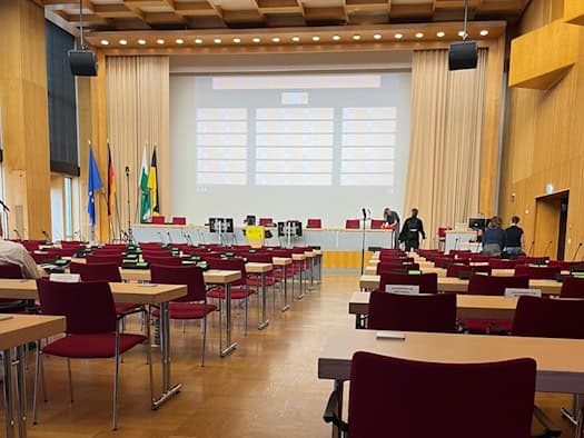 Blick durch die Reihen roter Stühle und Holztische im noch leeren Plenarsaal des Neuen Rathauses Dresden auf das Podium mit großer Leinwand und der Dresdner sowie sächsischen Flagge.