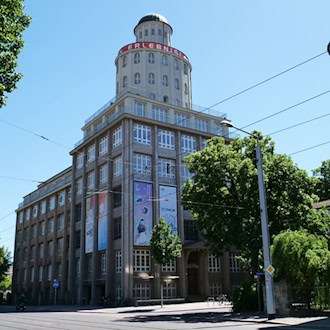 Außenansicht des historischen Ernemannbaus in Dresden bei sonnigem Wetter. Das mehrstöckige Industriegebäude der Technischen Sammlungen besitzt eine große Fensterfront und einen markanten runden Turm mit roter Aufschrift auf dem Dach.