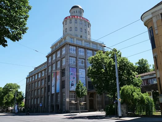 Außenansicht des historischen Ernemannbaus in Dresden bei sonnigem Wetter. Das mehrstöckige Industriegebäude der Technischen Sammlungen besitzt eine große Fensterfront und einen markanten runden Turm mit roter Aufschrift auf dem Dach.