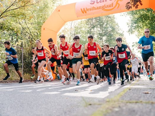 Eine Gruppe von Läuferinnen und Läufern, darunter Jugendliche in roten Trikots mit Startnummern, rennt unter einem großen orangen aufblasbaren Zielbogen des Westhanglaufs hindurch auf einer asphaltierten Straße im Park.